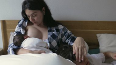 Anne yeni doğmuş bebeği emziriyor ve büyük çocuğu teselli ediyor. Huzurlu ve sevgi dolu bir evde ailenin şefkatli ve şefkatli bağını gösteriyor.