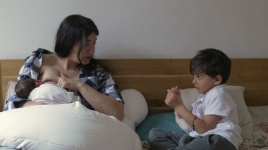 Anne bebeğini emziriyor, büyük oğluyla sohbet ediyor. Anne ekose gömlekli ve iki çocuk da yatağa yakın otururken aile yakınlaşması.