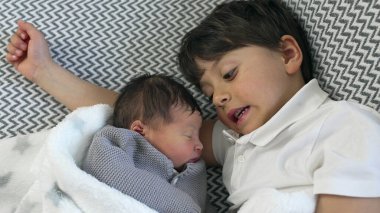 Küçük çocuk, yumuşak bir battaniyeye sarılı yeni doğmuş kardeşinin yanında yatar. Huzurlu ve samimi bir kardeş sevgisi ve aile bağını yakalar.