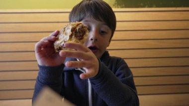 Genç çocuk büyük bir hevesle çizburgeri ısırıyor, elleri susamlı ekmeği tutuyor. İfade zevk ve açlık gösteriyor, lezzetli bir yemek yemenin basit zevkini vurguluyor.