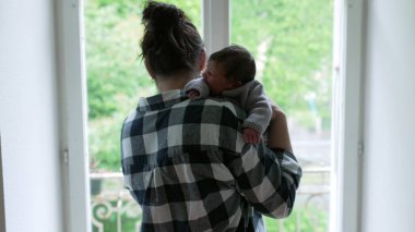 Anne, yeni doğmuş bebeğini omzuna almış pencerenin kenarında duruyor. Huzurlu bir bağ ve şefkat anı yakalıyor.