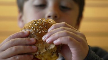 Çocuk şevkle sulu bir çizburgerden büyük bir ısırık alıyor, keyifli ve neşeli bir yüz ifadesi sergiliyor ve sıradan bir lokantada yemeğinin tadını çıkarıyor.