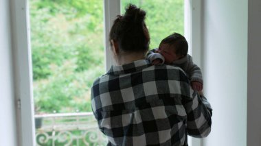 Anne, yeni doğmuş bebeğini omzuna almış pencerenin kenarında duruyor. Huzurlu bir bağ ve şefkat anı yakalıyor.