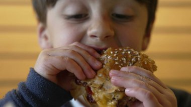 Çocuk şevkle sulu bir çizburgerden büyük bir ısırık alıyor, keyifli ve neşeli bir yüz ifadesi sergiliyor ve sıradan bir lokantada yemeğinin tadını çıkarıyor.