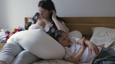 Anne, yeni doğan bebeği emzirirken büyük çocuğu teselli eder, huzurlu bir yuva ortamında bağ ve bakımı ön plana çıkarır, aile sevgisini ve desteğini gösterir.