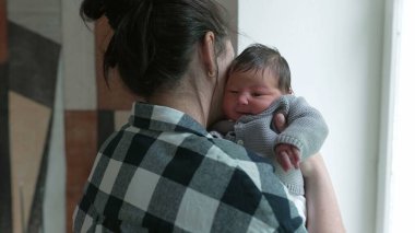 Anne pencerenin kenarında duruyor, omzunda yeni doğmuş bir bebekle, huzurlu bir bağ kurma anını yakalıyor.