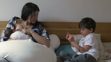 Anne bebeğini emziriyor, büyük oğluyla sohbet ediyor. Anne ekose gömlekli ve iki çocuk da yatağa yakın otururken aile yakınlaşması.