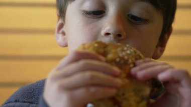 Çocuk şevkle sulu bir çizburgerden büyük bir ısırık alıyor, keyifli ve neşeli bir yüz ifadesi sergiliyor ve sıradan bir lokantada yemeğinin tadını çıkarıyor.