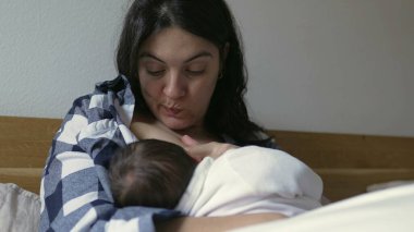 Anne, yeni doğmuş bebeği yatakta kareli gömlek giyerek nazikçe emziriyor. Beyaz bir battaniyeye sarılı bebek, barışçıl bir bağ anı paylaşıyor. Yakın çekim bağlantılarını yakalar