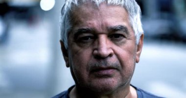 Gri saçlı ve ciddi ifadeli yaşlı adam şehir ortamında duruyor, geçmişi bulanık, yakın plan portresi, yüz detaylarını ve ifadelerini gösteriyor.