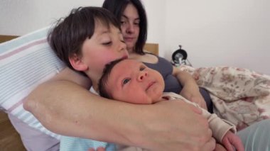 Anne ve büyük kardeş bebeği yatakta bir arada tutarlar, sevgi dolu bir aile anını yansıtırlar. Sahne aile üyeleri arasındaki samimiyet ve bağlantıyı yakalıyor, paylaştıkları bağ ve ilgiyi vurguluyor
