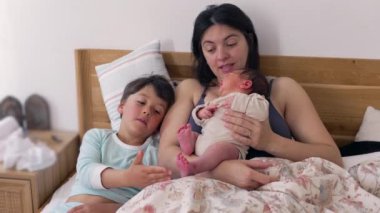 Anne bebeği tutarken, kardeş bebeğin ayağına hafifçe dokunuyor. Samimi aile anı, kardeşlerle anneleri arasında sıcak bir yuva ortamında paylaşılan sevgi ve bağın göstergesidir.