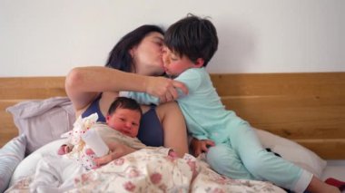 Anne, yeni doğmuş bebeği ve büyük çocuğuyla yatakta, samimi aile anı, kardeş şefkati, sıcak ve hassas atmosfer, yakın çekim, sevgi, bağ ve memnuniyeti vurguluyor.