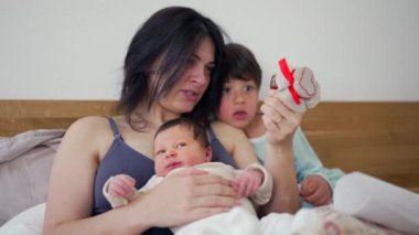 Anne, yeni doğmuş bebeğini kucağında tutarken yürekten bir hediye gösteriyor. Merakla izliyor, samimi bir aile anı. Sevgiye, kaynaşmaya, sıcaklığa ve ev yapımı hediyeye vurgu yapıyor.