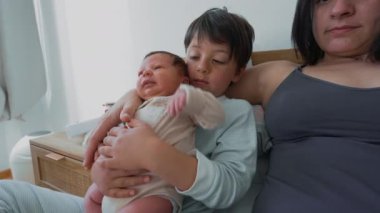 Anne, büyük kardeş merakla bakarken bebeği kucağına alır ve tatlı bir aile anını yakalar. Sahne anne ve çocuklar arasındaki derin bağ ve sevgiyi yansıtıyor, sıcaklık ve sevgiyle dolu.