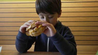 Lokanta ortamında ciddi bir ifadeyle lezzetli bir çizburgerin tadını çıkaran genç bir çocuk, rahat bir ortamda ahşap dekorlu ve doğal ışıklandırmalı konforlu yemekler yemenin sevincini sergiliyor.