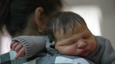 Anne, huzurlu, güneşli bir odada bebeğini nazikçe omzunda tutuyor. Bebek mışıl mışıl uyuyor, anne ve çocuk arasındaki derin sevgiyi ve şefkati gösteriyor.