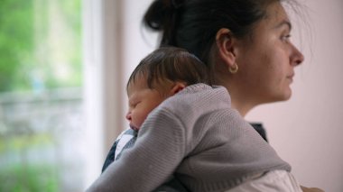 Anne, yeni doğmuş bebeğini omzunda beşikte, güneşli pencereden dışarı bakıyor. Sakin ve besleyici bir ortamda anne ile çocuk arasında sakin ve samimi bir bağ.