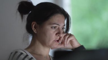 Evde dizüstü bilgisayarı üzerinde çalışan bir kadının odaklanmış ifadesine yakın çekim yapmak, bir iç ofis ortamında uzaktan çalışmanın yoğunluğunu ve adanmışlığını vurgulamak, kişisel ve iş yaşamını harmanlamak,