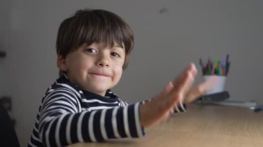 Genç bir çocuk, neşeli bir gülümsemeyle elini sallıyor, çizgili bir tişört giymiş, tahta bir masada oturuyor, neşeli ve neşeli bir çocukluk anını yakalıyor.