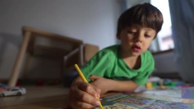 Yeşil tişörtlü çocuk yerde sarı işaretli resim çiziyor, ev ortamında rahat, boyama kitabına odaklanmış yaratıcı etkinlik, doğal ışık, kapalı mekanda, çocukluk eğlencesi...