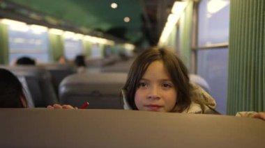 Elinde kırmızı bir kalemle tren koltuğuna bakan genç bir kız, yumuşak yeşil bir iç mekan ile çevrili ve arka planda bulanık yolcular...
