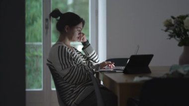 Evde dizüstü bilgisayarda çalışan, düşünceli bir ifadeyle yemek masasında oturan, uzaktan çalışma yaşam tarzını vurgulayan ve modern hayattaki profesyonel ve kişisel alanların karışımına dikkat çeken bir kadın.