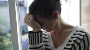 Yüzünü eliyle kapatan, pencere kenarında oturan, bunalmış hisseden bir kadın. Çizgili Süveter, Doğal Işık, İç mekan. Gerçek Hayat Duyguları, Stres, Endişe ve Kişisel Mücadeleyi Yakalar