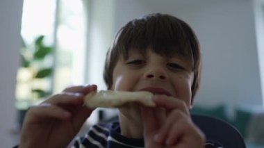 Lezzetli bir atıştırmalığı seven, peynirli bir parça ekmek tutan, çocukluk sevincini yakalayan, yemek yemenin basit zevklerini alan, arka planda bulanık ve yumuşak bir ışık alan genç bir çocuk.