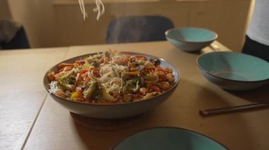 Elleri sebze ve karideslerle dolu canlı bir erişte kızartması servis ediyor, tabağı karıştırmak için turuncu tencereler kullanıyor, sıcak bir mutfakta ev yapımı Asya mutfağının özünü yakalıyor.