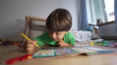 Sarı keçeli kalemle boyalı, rahat bir evde yerde yatıyor, yeşil tişört giyiyor, boyama kitabına odaklanmış, yaratıcı etkinlik, kapalı mekanda, doğal ışık, çocukluk gelişimi...