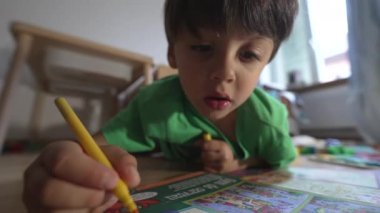 Yeşil tişörtlü çocuk yere sarı işaretli resim çiziyor, ev dekorasyonu, boyama kitabı, yaratıcı ve eğitici oyun, kapalı mekan, doğal ışık, çocukluk gelişimi üzerine odaklanıyor.