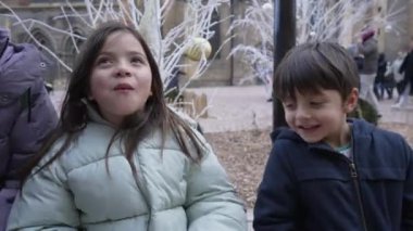 Neşeli çocuklar, açık hava festivalinde kış tatilinin tadını çıkarıyorlar. Gülümseyen çocuk ve kız, soğuk hava ve canlı atmosferi kucaklıyor, etrafı tatil dekorasyonlarıyla çevrili.