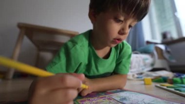 Sarı kalemle çizim yapan, rahat ev ortamında yerde yatan, yeşil gömlek giyen, boyama kitabına odaklanan, kapalı alanda yaratıcı ve eğitici oyun oynayan bir çocuk.