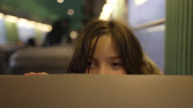 Kabarık bir kışın elinde kırmızı bir kalemle gülen kız, yeşil iç mekânlı tren koltuğunda pena-boo oynuyor, arka planda parlak ışıklar yanıyor ve bulanık şekiller, sıcak ve mutlu bir atmosfer yaratıyor.