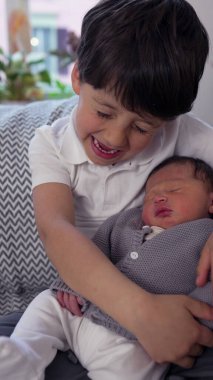 Genç çocuk, yeni doğmuş kardeşini kanepede kucaklıyor, ev dekoruyla çevrili, yakın bağlarını ve kardeş sevgisini vurguluyor.