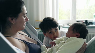 Anne ve büyük kardeş hastane yatağında yeni doğan bebeğe hayran. Sevecen bir etkileşim ailenin neşesini ve kardeşler arasındaki yakın bağı vurgular. Yürek ısıtan bir sevgi ve bağ anını yakalar.