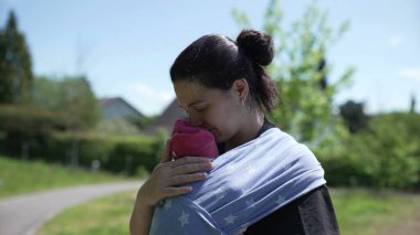 Anne, yeni doğmuş bebeğini nazikçe sarmalayıp yeşil ağaçlar ve açık gökyüzü olan bir parkta duruyor. Erken ebeveynlik ve açık hava ilişkisinin dingin ve besleyici anlarını vurguluyor.