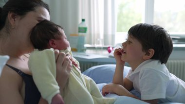Anne ve küçük oğlu bir hastane odasında yeni doğan bebeğe hayran. Genç çocuğun gülümsemesi ve annesinin şefkatli bakışları bir ailenin yeni üyesini karşılama sevincini ve heyecanını gösteriyor.
