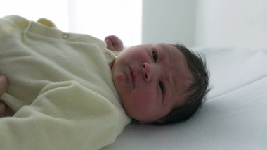 Hemşire onu hastanede giydirip düğmelerini iliklerken yeni doğmuş bebek sakin ve huzurlu. Bebek yüzündeki sakin ifade, rutin bebek bakımı sırasında hemşirenin sağladığı nazik ve hassas bakımı vurgular.