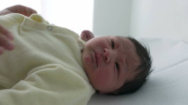 Hemşire onu hastanede giydirip düğmelerini iliklerken yeni doğmuş bebek sakin ve huzurlu. Bebek yüzlerindeki sakin ifade, hemşirenin rutin bebek bakımı sırasında gösterdiği nazik ve hassas ilgiyi vurgular. 