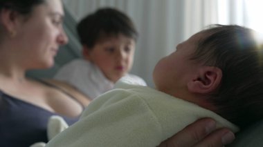 Anne ve büyük kardeş yeni doğan bebeğe bakıyor, aile içi sevgi ve hastane yatağında bir bağ yakalıyorlar. Anne, büyük çocuk ve yeni doğan arasındaki hassas etkileşim ve bağ