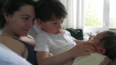 Anne ve büyük kardeş hastane yatağında yeni doğan bebeğe nazikçe hayran. Aile anlarında kardeşlerin sıcaklığı ve bağlantıları vurgulanıyor. Anne şefkati, aile neşesi ve birlikteliği vurgulanıyor.