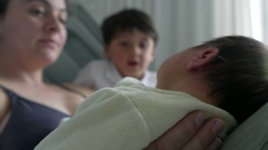 Anne ve büyük kardeş yeni doğmuş bebeğe sarılıyor, sıcak bir şekilde gülümsüyor. Samimi aile anları, anne ve çocuklar arasındaki sevgi ve bağı yakalar. İlişkilerindeki neşe ve bağlantıyı vurgular.