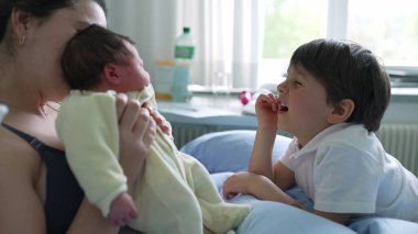 Anne ve küçük oğlu bir hastane odasında yeni doğan bebeğe hayran. Genç çocuğun gülümsemesi ve annesinin şefkatli bakışları bir ailenin yeni üyesini karşılama sevincini ve heyecanını gösteriyor.