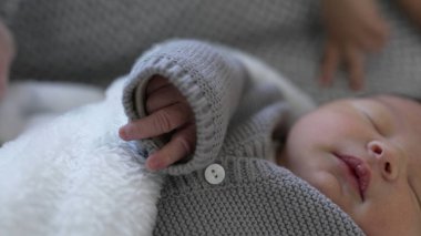 Yeni doğmuş bebek örgülü gri bir elbiseyle huzur içinde uyuyor. Kolundan küçük bir el çıkıyor. Yumuşak, rahat bir battaniyeye sarılmış.