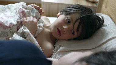 Çocuk battaniyenin altında yatakta yatıyor, sabah rutini, rahat yatak odası, uyanıyor, günün rahat başlangıcı, aile anı, çocuk bakıyor, rahat yatak örtüsü, sabah ışığı