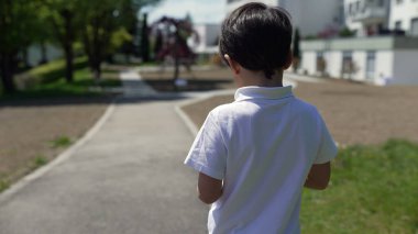 Bir yolun üzerinde duran, çocuk parkına ve konut binalarına bakan, güvenli, banliyö ortamında merak ve keşifleri ön plana çıkaran genç bir çocuğun arka görüntüsü