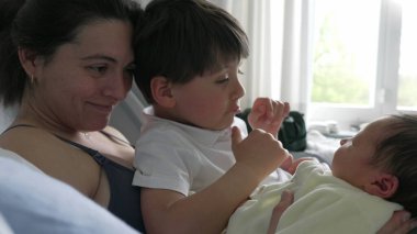 Anne ve büyük kardeş hastane yatağında yeni doğan bebeğe nazikçe hayran. Aile anlarında kardeşlerin sıcaklığı ve bağlantıları vurgulanıyor. Anne şefkati, aile neşesi ve birlikteliği vurgulanıyor.