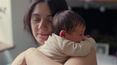 Anne yeni doğmuş bebeği kucaklar, gözleri saf şefkat dolu bir anda kapanır. Samimi ortam ve nazik ışık anne ve çocuk arasındaki derin bağ ve sevgiyi vurguluyor.